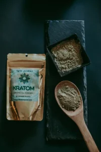 Top view of kratom powder in a bowl and spoon, alongside a branded package, on a dark surface.