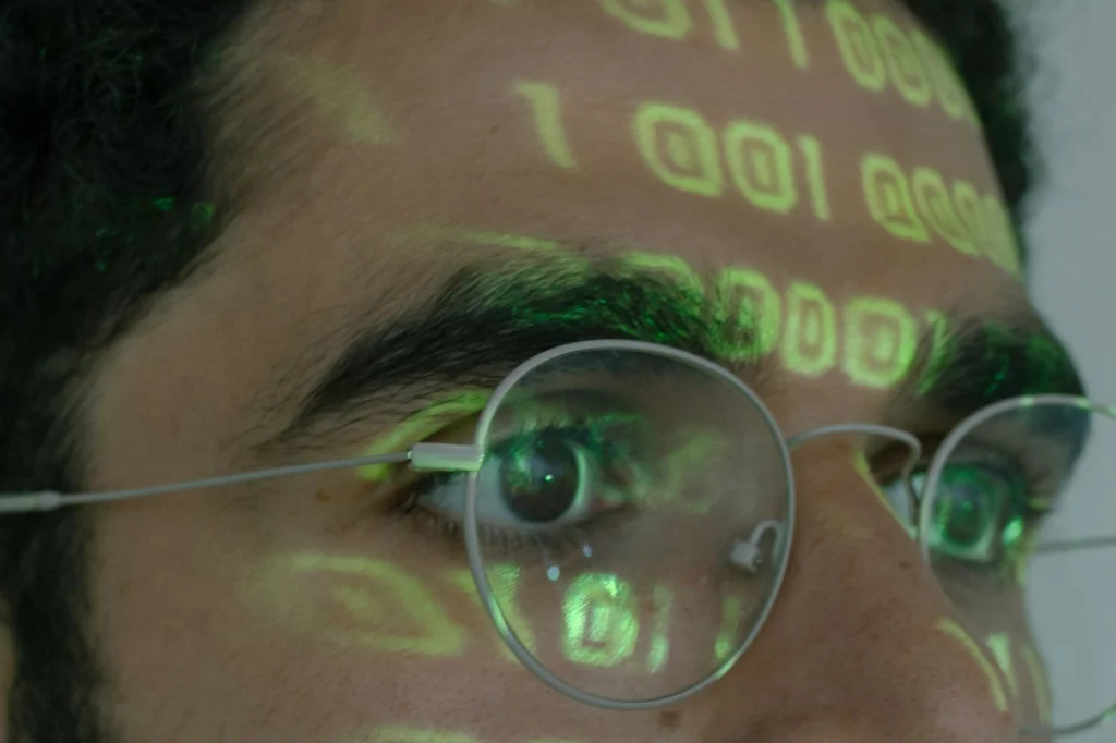 Detailní snímek osoby, na jejíž brýle je promítán binární kód, symbolizující digitální technologie.