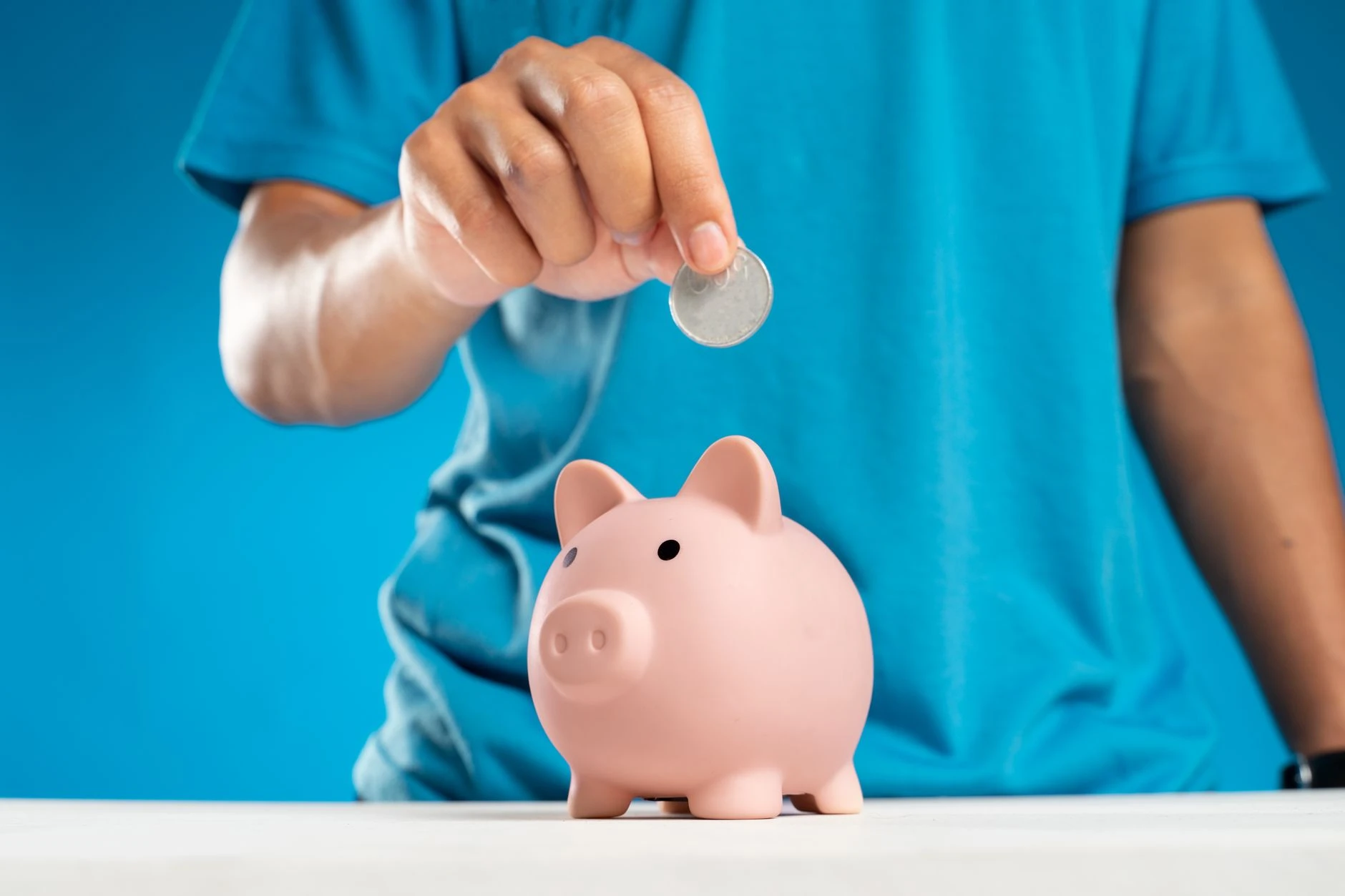 Detailní záběr na ruku dospělého, která vkládá minci do prasátka, symbolizující spoření a investice.