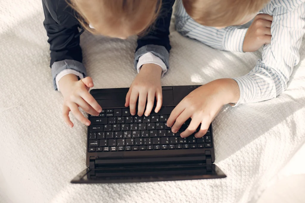 Dvě děti ve volném oblečení sdílejí notebook na útulné posteli, podporující týmovou práci a online učení.