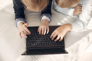 Dvě děti ve volném oblečení sdílejí notebook na útulné posteli, podporující týmovou práci a online učení.