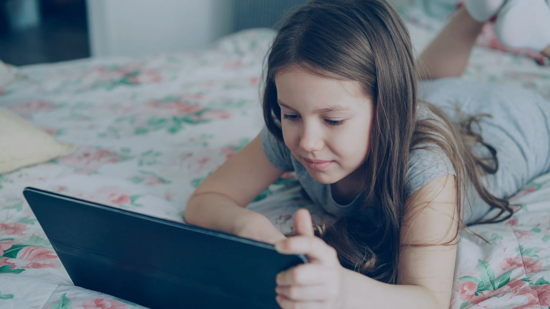 Mladá dívka leží na posteli a používá tablet, ponořená do svého digitálního světa.