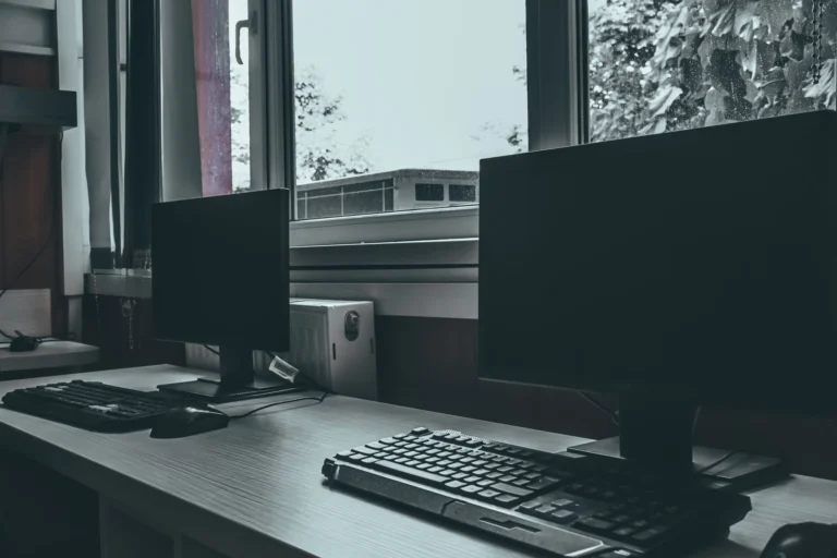 Prázdné kancelářské pracovní místo se dvěma počítačovými monitory u okna, vyjadřující moderní a profesionální prostředí.
