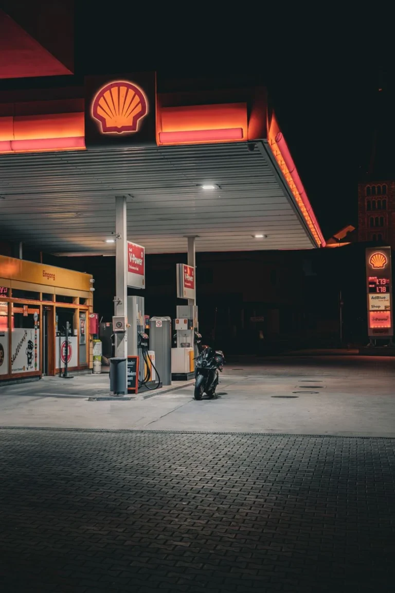 Slabě osvětlená benzínka Shell s motocyklem zaparkovaným pod jasnými světly během noci.