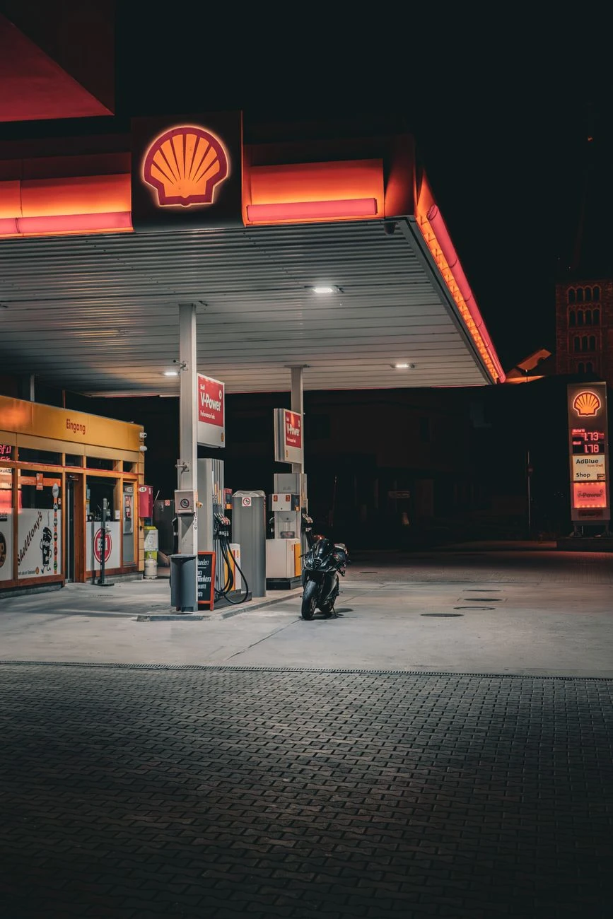 Slabě osvětlená benzínka Shell s motocyklem zaparkovaným pod jasnými světly během noci.