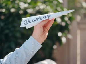 Detailní záběr na ruku, která drží papírovou loďku s nápisem 'Startup', symbolizující zahájení podnikání.