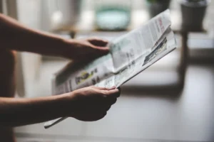 Ruce držící noviny v měkkém osvětlení vnitřního prostředí, soustředěné na uvolněný okamžik čtení.